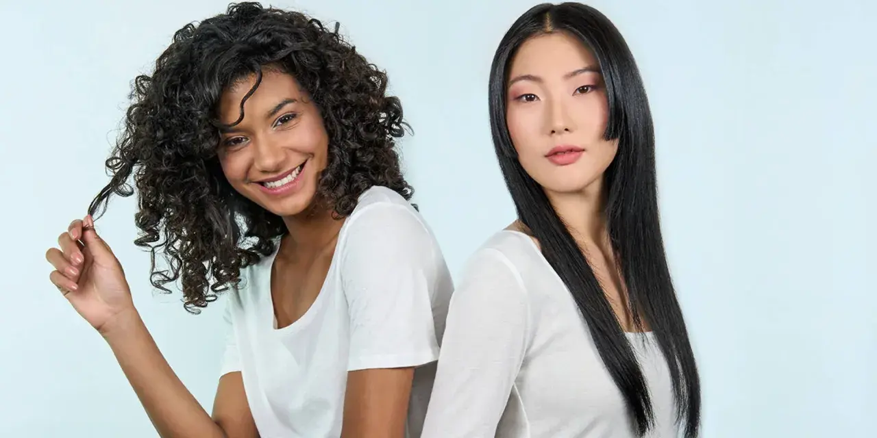Deux femmes souriantes, l'une aux cheveux bouclés, l'autre aux cheveux lisses. La première joue avec ses mèches, évitant les cheveux fourchus jusqu'à la racine.