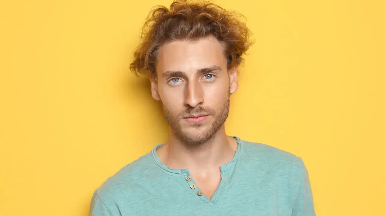 Un jeune homme aux cheveux bouclés et aux yeux bleus regarde la caméra sur fond jaune.