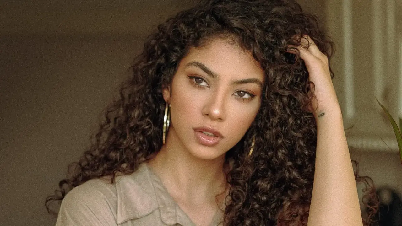 Une jeune femme aux cheveux bruns et bouclés, portant une chemise beige et de grandes boucles d'oreilles. Sa coupe boucles met en valeur son regard intense.