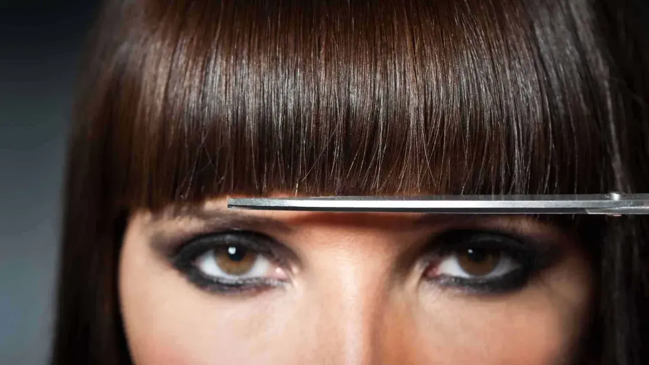 Un modèle de coiffure aux yeux marron et au maquillage prononcé, une frange brune est coupée par des ciseaux.