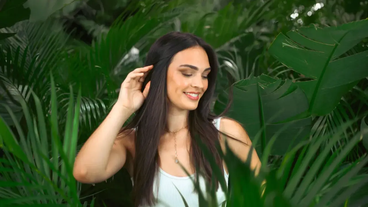 Une jeune femme aux cheveux lisses et sombres, souriant doucement, se tient parmi des feuilles vertes luxuriantes.