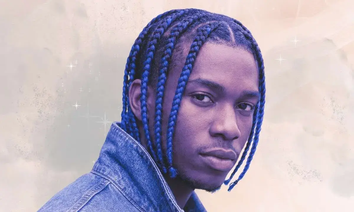 Un jeune homme aux tresses bleues éclatantes, portant une veste en jean, pose avec confiance.