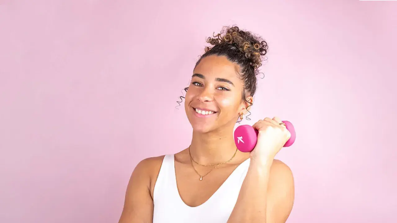 Une jeune femme souriante avec une coiffure sport cheveux bouclés tient une haltère rose.