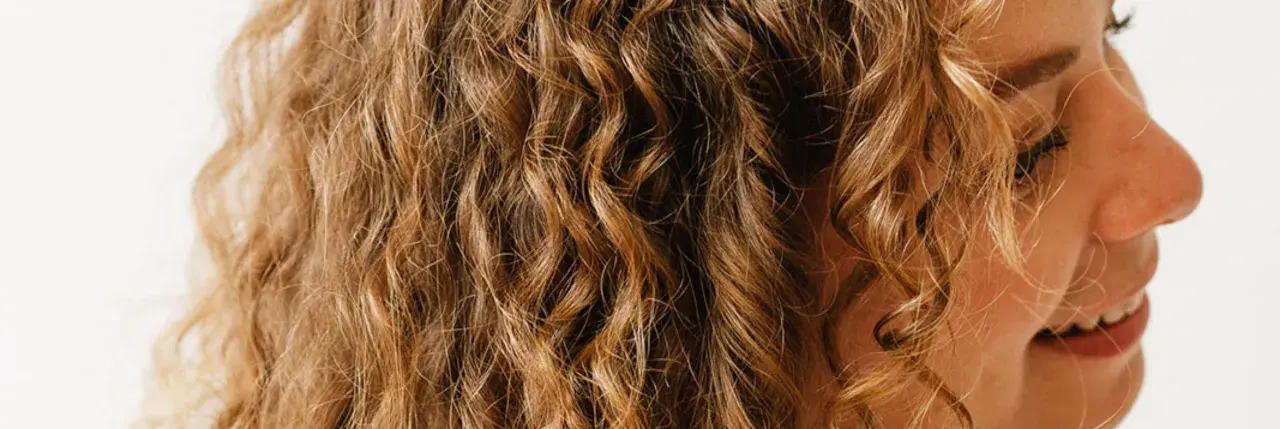 Une femme se coiffe les cheveux ondulés le matin, révélant des boucles dorées et un sourire radieux.