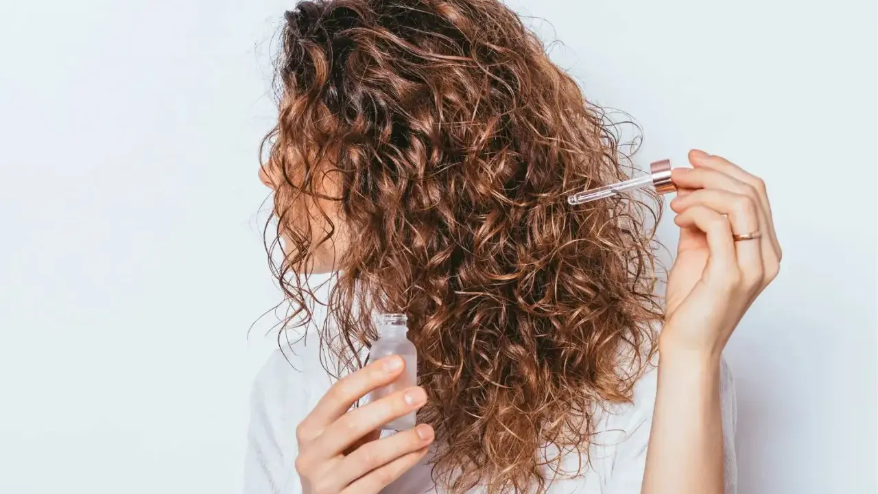 Une femme applique une huile capillaire sur ses cheveux frisottis sur le dessus. Ses boucles brunes brillent sous la lumière.