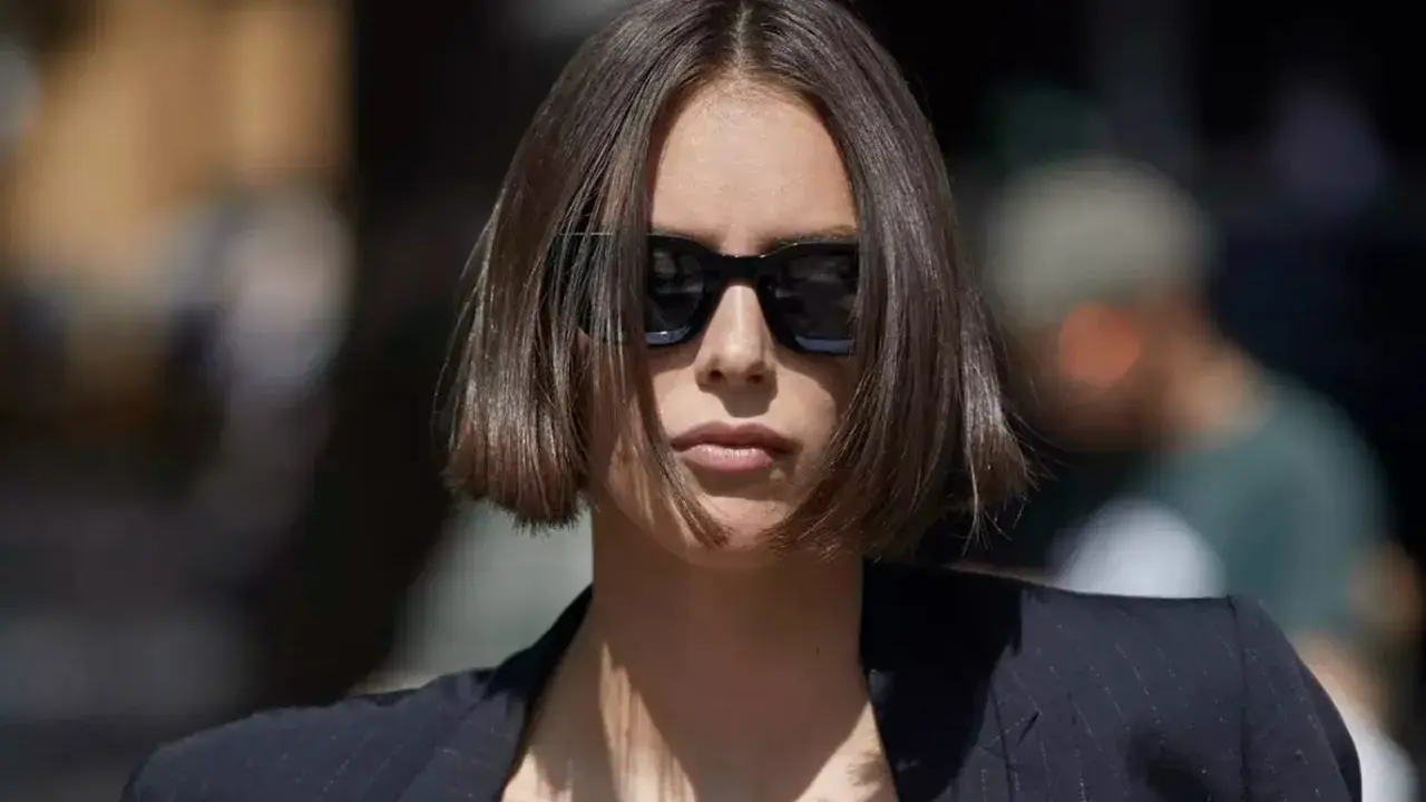 Une femme au regard intense arbore une coupe carré droit dégradé, accentuée par des lunettes de soleil noires et un blazer chic.