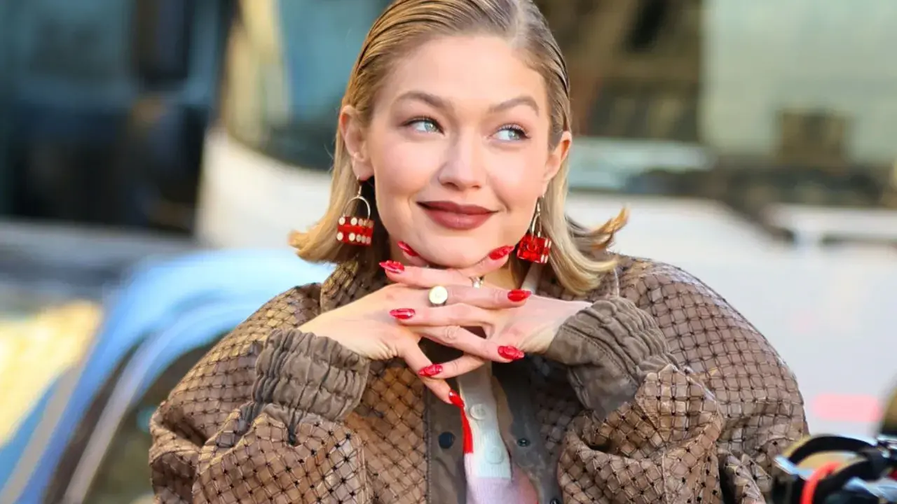 Gigi Hadid, avec ses boucles d'oreilles en forme de dé rouge, pose avec un sourire malicieux. Son carré plaqué en arrière et son manteau texturé complètent son look.