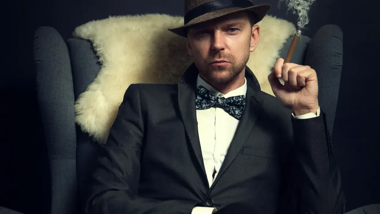 Homme au costume élégant, chapeau et nœud papillon, avec une coupe cheveux rockabilly homme. Il fume un cigare, assis dans un fauteuil.