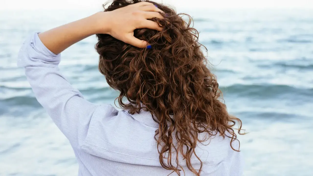 Main tenant ses cheveux bouclés, une femme en chemise rayée profite de la brise marine. Elle n'a pas peur de brosser ses cheveux bouclés tous les jours.