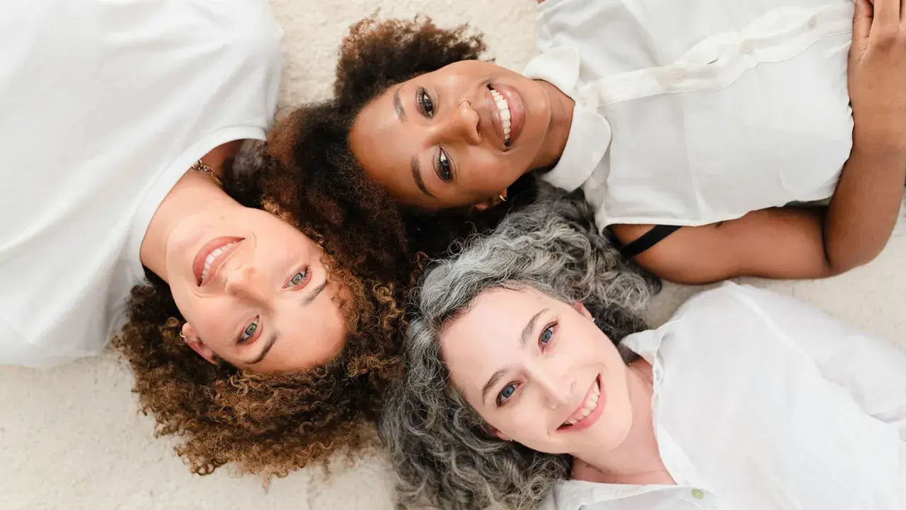 Trois femmes souriantes, aux cheveux bouclés, frisés et gris, partagent un moment complice. Un scruncher cheveux pourrait parfaire leur look.