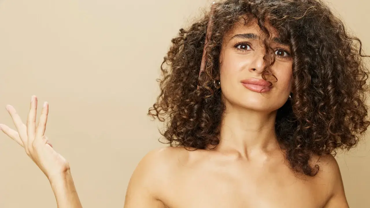 Une femme aux cheveux longs et bouclés, une brosse à cheveux coincée dedans, lève une main ouverte.