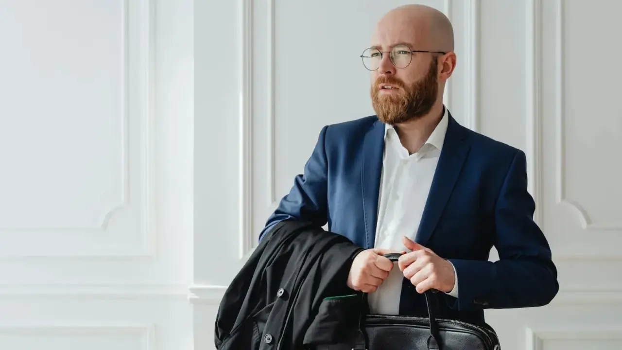 Un homme crane rasé, barbe rousse, lunettes, en costume bleu, porte une veste noire et une mallette.