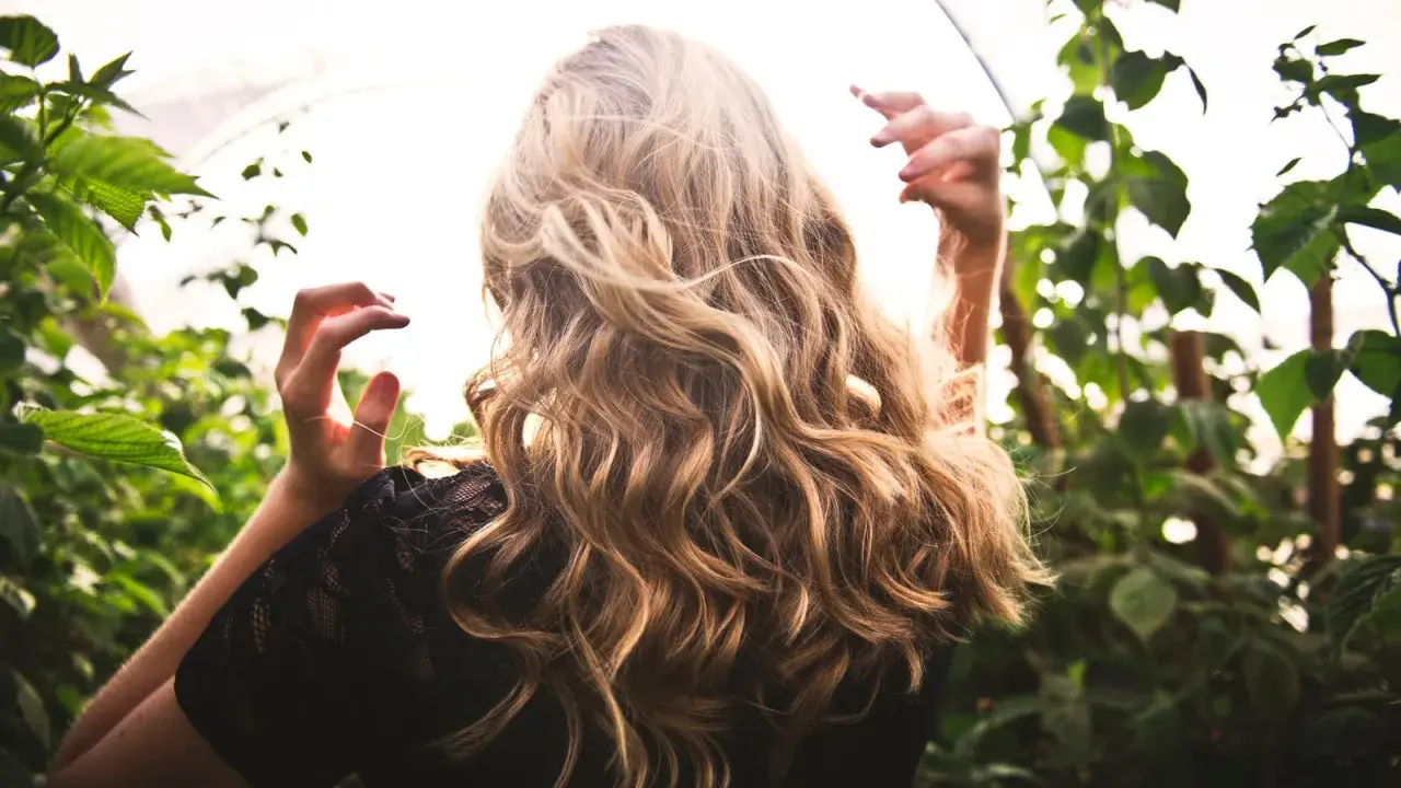 Chevelure blonde ondulée, baignée de lumière, entourée de verdure. Une scène évoquant la douceur d'une **décoloration végétale** naturelle.