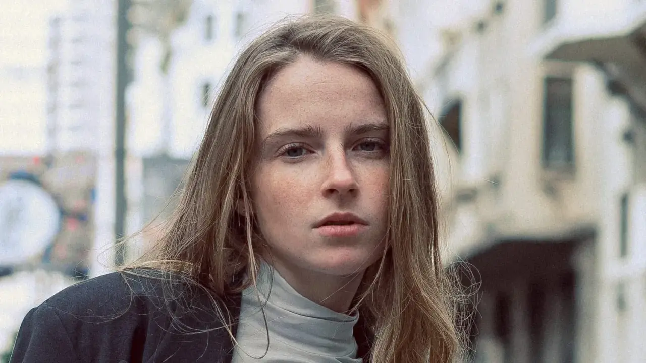 Jeune femme au regard intense, ses cheveux châtain clair cendré ou glacé encadrent son visage parsemé de taches de rousseur. Elle porte un col roulé gris et une veste sombre.