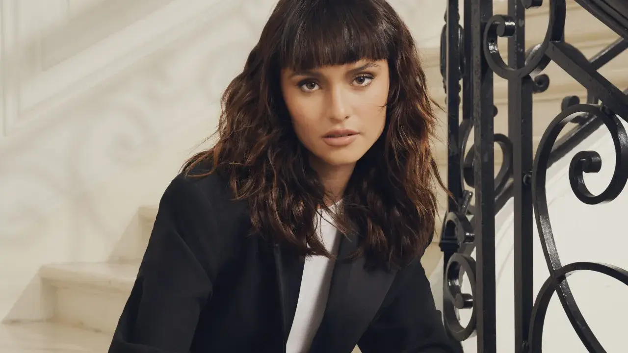 Une femme au regard intense arbore un **wolf cut cheveux bouclés** brun, porté avec un blazer noir.