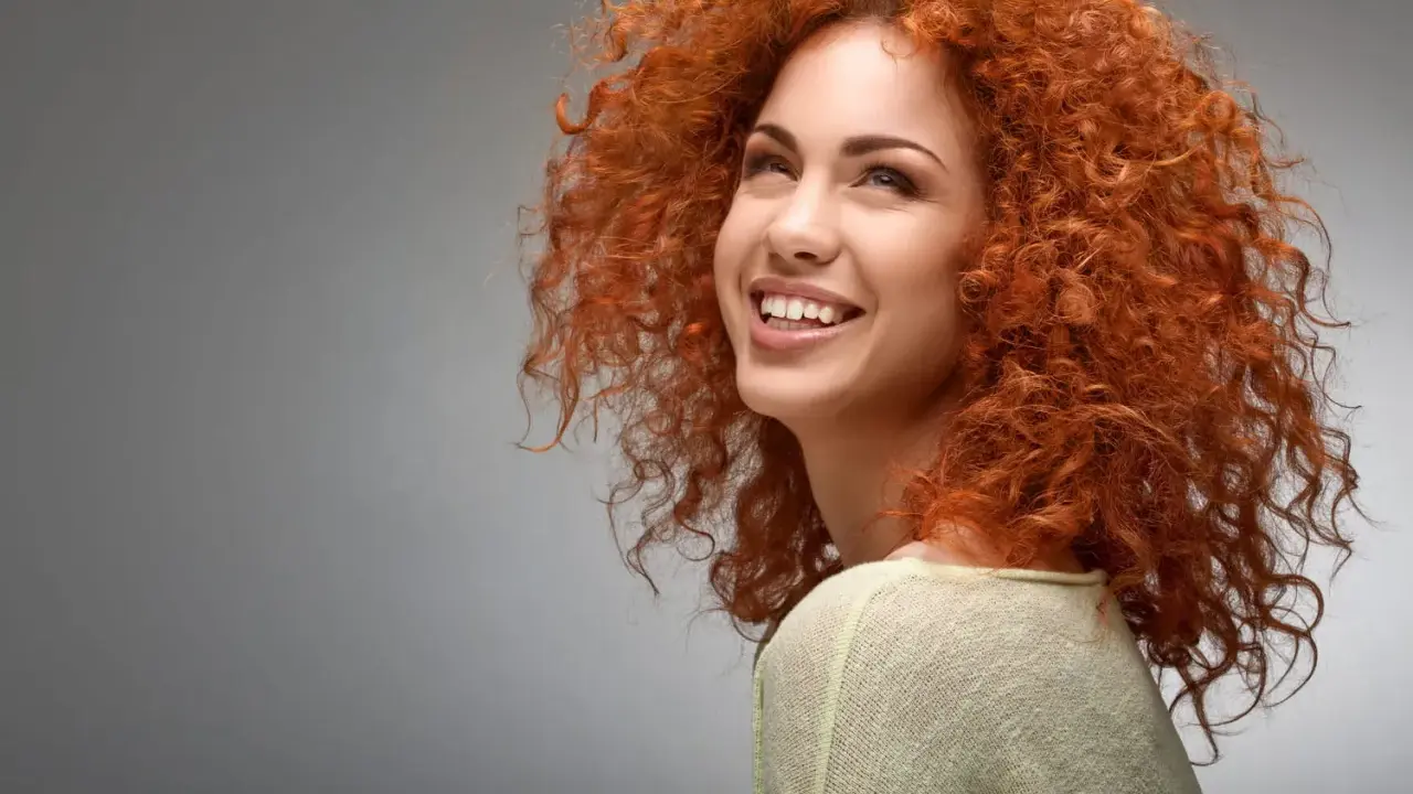 Une jeune femme au dégradé cheveux bouclés mi long, rousse, sourit joyeusement.