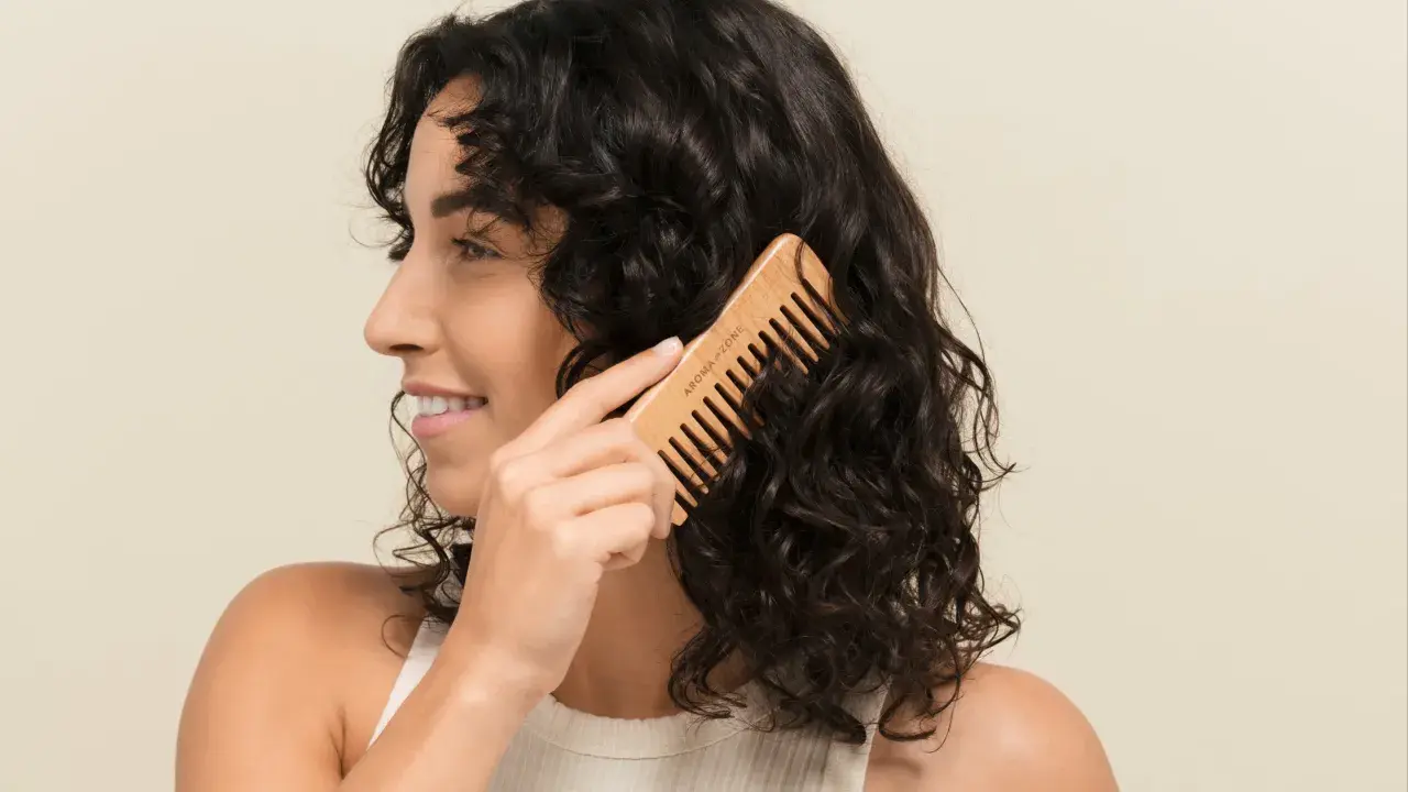Une femme se peigne les cheveux bruns avec un peigne en bois. Elle sourit, car mes cheveux ne bouclent pas partout, mais ce peigne aide à définir les boucles.