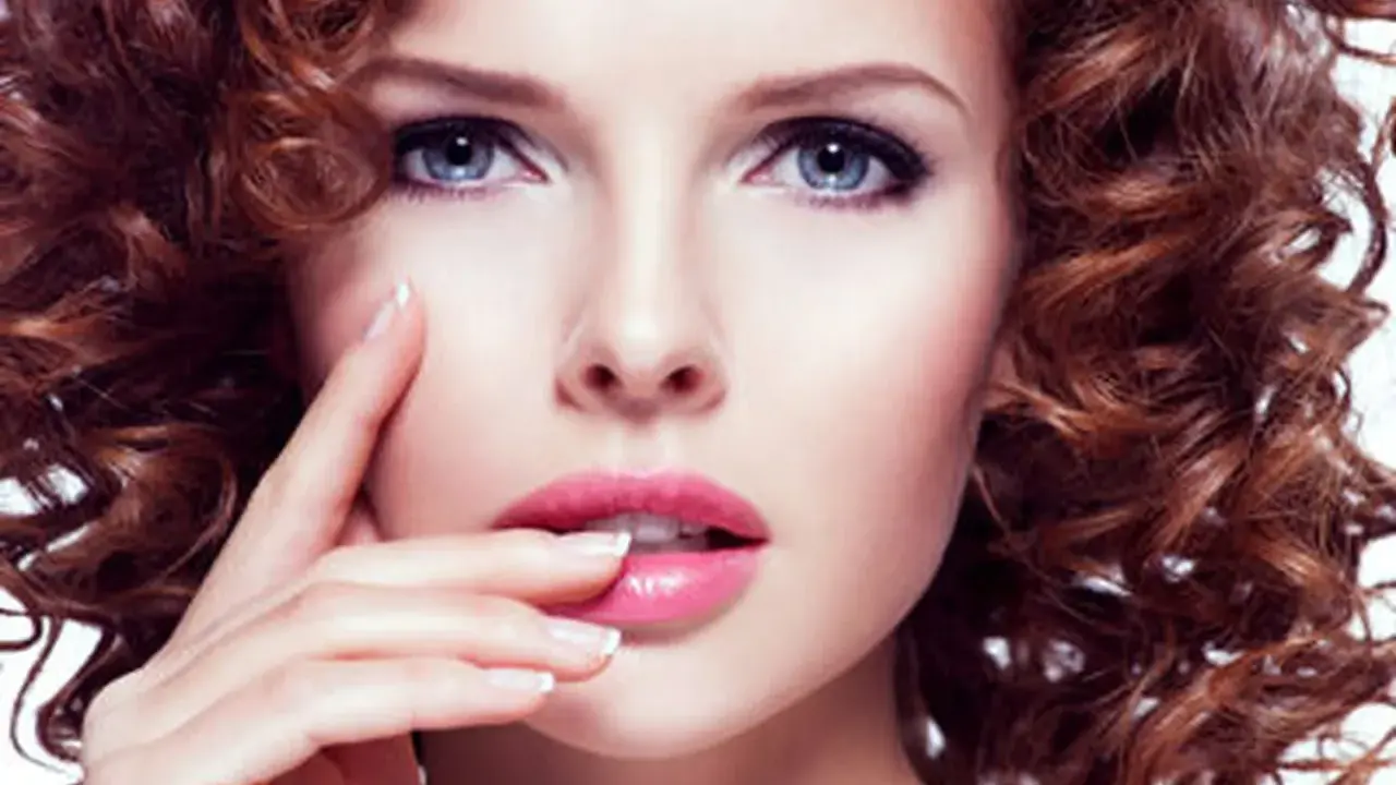 Une femme à la peau claire et aux yeux bleus, avec une chevelure rousse bouclée. Ses lèvres sont roses et brillantes.