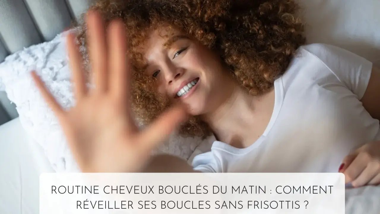 Une femme souriante aux cheveux 3c bouclés se réveille dans son lit. Sa main est devant l'objectif.