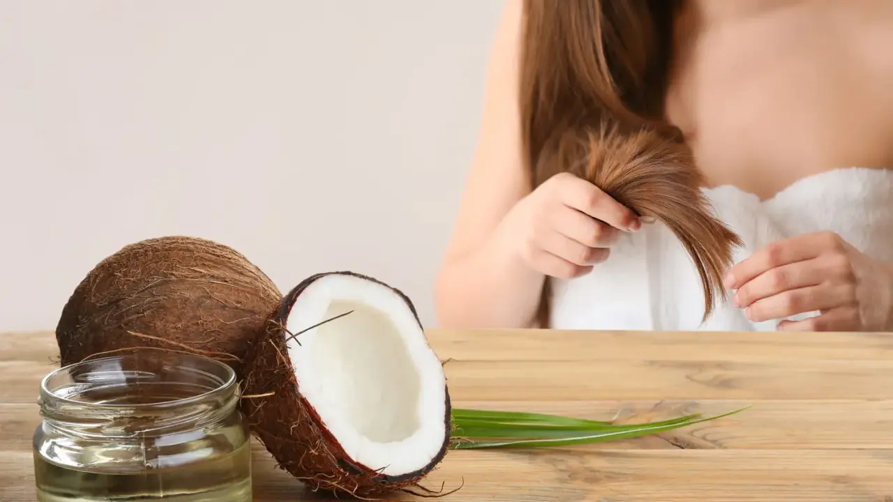 Un pot d'huile de coco et des noix de coco sur une table en bois, avec une femme en arrière-plan qui touche ses cheveux. L'huile de coco est idéale pour les cheveux bouclés.