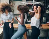 Trois femmes aux cheveux bouclés dansent joyeusement. Leurs chevelures volent, un exemple de comment avoir de belles boucles sans frisottis.
