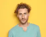 Un jeune homme aux cheveux bouclés et aux yeux bleus regarde la caméra sur fond jaune.