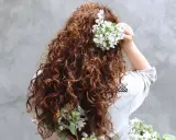 Chevelure brune et bouclée ornée de fleurs blanches, évoquant un décollement de racine naturel et printanier.