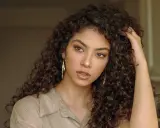 Une jeune femme aux cheveux bruns et bouclés, portant une chemise beige et de grandes boucles d'oreilles. Sa coupe boucles met en valeur son regard intense.