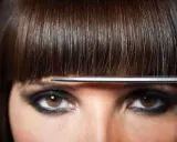 Un modèle de coiffure aux yeux marron et au maquillage prononcé, une frange brune est coupée par des ciseaux.