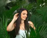 Une jeune femme aux cheveux lisses et sombres, souriant doucement, se tient parmi des feuilles vertes luxuriantes.