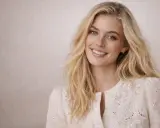Une jeune femme au sourire radieux, ses **cheveux décolorés** tombant en cascade sur ses épaules, porte une blouse blanche brodée.