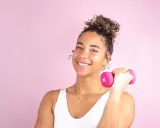 Une jeune femme souriante avec une coiffure sport cheveux bouclés tient une haltère rose.
