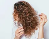 Une femme applique une huile capillaire sur ses cheveux frisottis sur le dessus. Ses boucles brunes brillent sous la lumière.