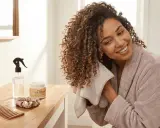 Une femme souriante utilise une serviette sur ses cheveux bouclés. Sur la table, un peigne, une crème pour boucles et un spray d'eau pour un tuto coiffure cheveux bouclés naturel.