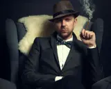 Homme au costume élégant, chapeau et nœud papillon, avec une coupe cheveux rockabilly homme. Il fume un cigare, assis dans un fauteuil.