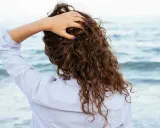 Main tenant ses cheveux bouclés, une femme en chemise rayée profite de la brise marine. Elle n'a pas peur de brosser ses cheveux bouclés tous les jours.