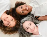 Trois femmes souriantes, aux cheveux bouclés, frisés et gris, partagent un moment complice. Un scruncher cheveux pourrait parfaire leur look.
