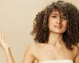 Une femme aux cheveux longs et bouclés, une brosse à cheveux coincée dedans, lève une main ouverte.