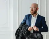 Un homme crane rasé, barbe rousse, lunettes, en costume bleu, porte une veste noire et une mallette.