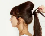 Une main ajuste une mèche pour créer un chignon sophistiqué. La coiffure volumineuse est en cours de réalisation.