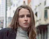 Jeune femme au regard intense, ses cheveux châtain clair cendré ou glacé encadrent son visage parsemé de taches de rousseur. Elle porte un col roulé gris et une veste sombre.
