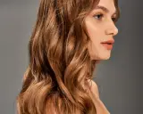 Chevelure ondulée, passant du blond au chatain, d'une femme au profil délicat, sur fond gris.
