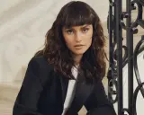 Une femme au regard intense arbore un **wolf cut cheveux bouclés** brun, porté avec un blazer noir.