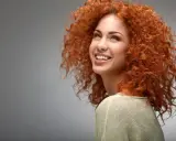 Une jeune femme au dégradé cheveux bouclés mi long, rousse, sourit joyeusement.