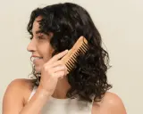 Une femme se peigne les cheveux bruns avec un peigne en bois. Elle sourit, car mes cheveux ne bouclent pas partout, mais ce peigne aide à définir les boucles.