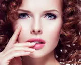Une femme à la peau claire et aux yeux bleus, avec une chevelure rousse bouclée. Ses lèvres sont roses et brillantes.