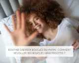 Une femme souriante aux cheveux 3c bouclés se réveille dans son lit. Sa main est devant l'objectif.