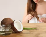 Un pot d'huile de coco et des noix de coco sur une table en bois, avec une femme en arrière-plan qui touche ses cheveux. L'huile de coco est idéale pour les cheveux bouclés.