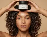 Une femme aux cheveux bouclés tient un pot de masque perfecteur de boucles au beurre de karité au-dessus de sa tête.