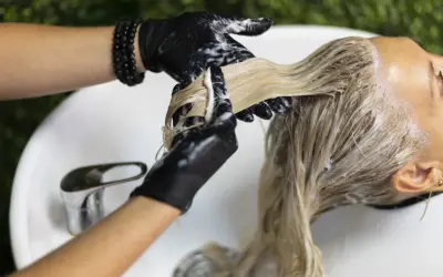 Application d'une patine sur des cheveux blonds. Le coiffeur, portant des gants noirs, applique le produit. Le temps de pose est crucial pour le résultat.